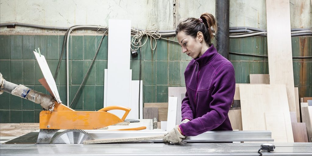 Woman in a purple jacket and gloves cutting lumber on a table saw in a wood shop