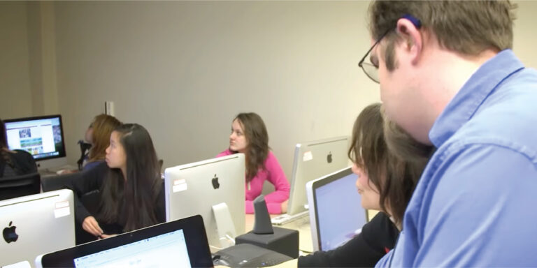 Students in a classroom using Macintosh computers looking up at a lecturer.