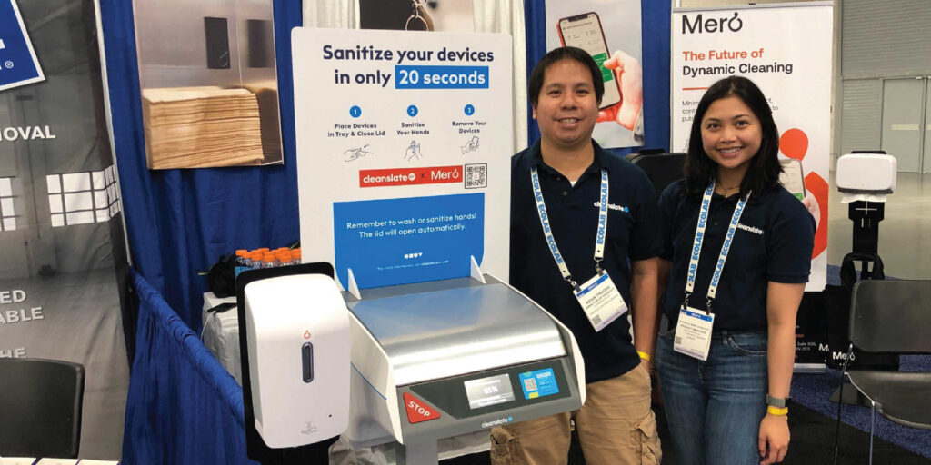Two people at a display showing the CleanSlate UV Mero device under a poster that says "Sanitizes your devices in only 20 seconds."