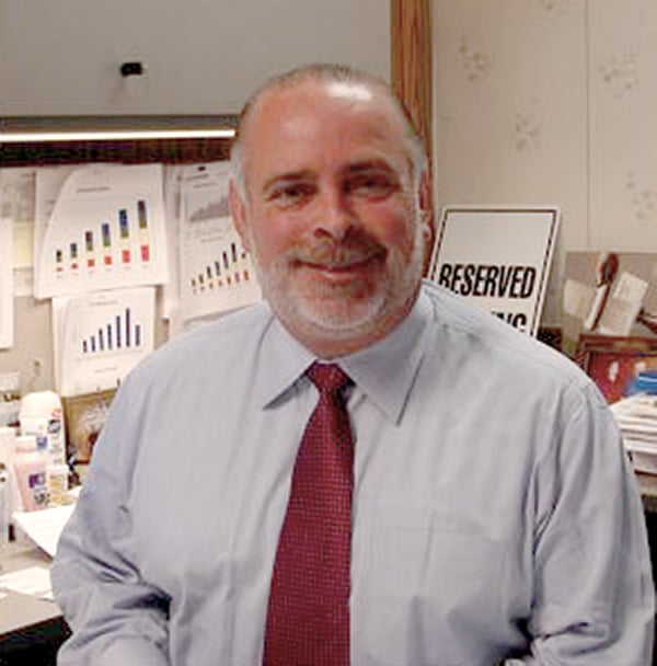 Dave Pelligrini at his desk in a shirt and tie with graphs and charts on a board behind him.